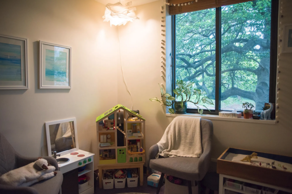 A counseling office illuminated by a hanging light, featuring a playhouse, chair, and sand tray. 